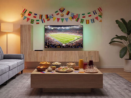 Woonkamer ingericht om naar een voetbalwedstrijd te kijken, met een tv waarop een wedstrijd in een stadion wordt uitgezonden, een bank tegenover een houten salontafel vol snacks en drankjes, en kleurrijke slingers met internationale vlaggen die de muur decoreren. Klik om de producten uit de collectie te bekijken.