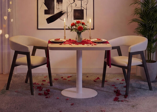 Romantische dineropstelling in een moderne woonkamer met een kleine ronde witte tafel en twee gestoffeerde stoelen, gedekt met borden, kaarsen, een vaas met rode rozen en verspreide rozenblaadjes op tafel en tapijt, omlijst door zachte gordijnverlichting, abstracte wandkunst en een groene kamerplant voor een gezellige Valentijnssfeer.