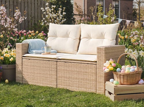 Een wicker tuinbank met twee zitplaatsen en cr&egrave;me kussens op een groen gazon, omringd door bloeiende voorjaarsbloemen, een mand met gekleurde paaseieren en feestelijke slingers. Klik om de producten in de collectie te bekijken.