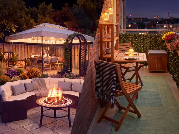 Knusse buitenruimtes in de schemering, met aan de ene kant een tuinloungeset met vuurplaats en een prieel vol bloemen, en aan de andere kant een ingericht balkon met houten meubels, lantaarnverlichting en bloemen in potten met uitzicht op een stadssilhouet.