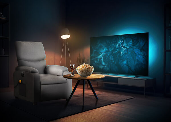 Gezellige thuisbioscoop-scène met een grijze fauteuil naast een houten salontafel met popcorn en frisdrank, een vloerlamp die zachtjes in de hoek straalt en een grote flatscreentelevisie met blauwe sfeerverlichting in een moderne woonkamer.