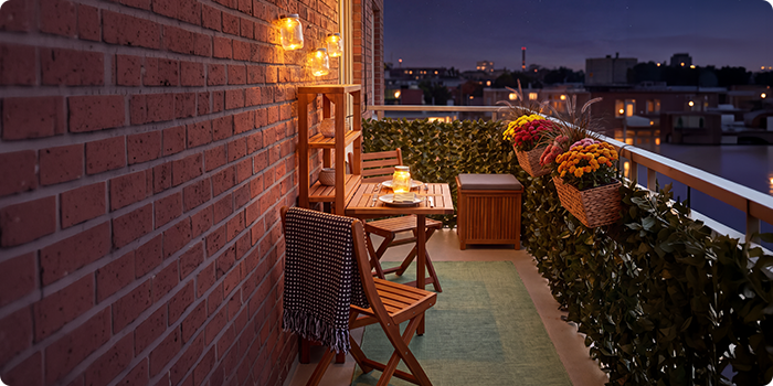 Klein balkon met houten stoelen en tafel, verlicht met lantaarns en kaarsen, versierd met bloemen in potten en groen, met uitzicht op een stadssilhouet bij nacht.