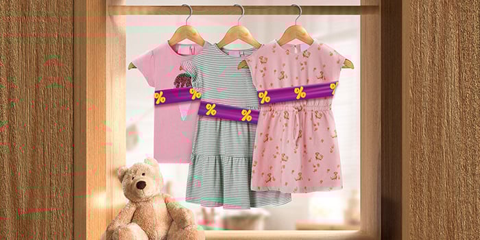 Kinderkleding op hangers aan een rail binnen, omwikkeld met een paarse promotieband met gele kortingspictogrammen. Klik om de producten in de aanbieding te bekijken.