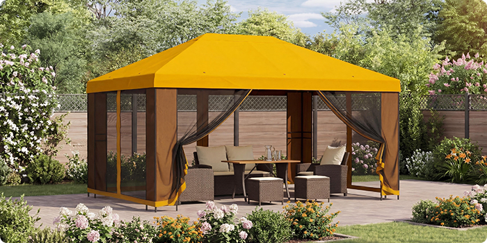 Gele tuinpaviljoen met gaasgordijnen dat een loungeset met tafel op een terras overdekt, omringd door bloeiende planten in een zonnige tuin.
